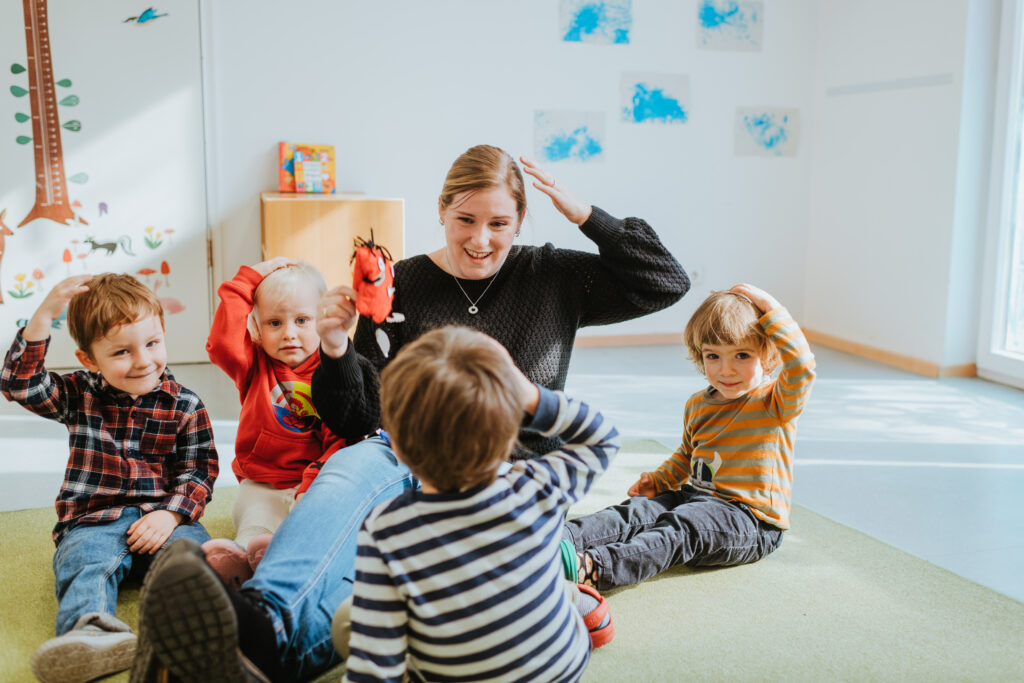 Eine junge Frau sitzt mit vier Kindern im Kreis auf einem grünen Teppich. Sie hält eine rote Handpuppe hoch und alle legen sich die Hände auf den Kopf, vermutlich im Rahmen eines Spiels oder Lieds. Die Kinder sind fröhlich und aufmerksam. Im Hintergrund ist ein heller Raum mit Kinderzeichnungen an der Wand zu sehen.