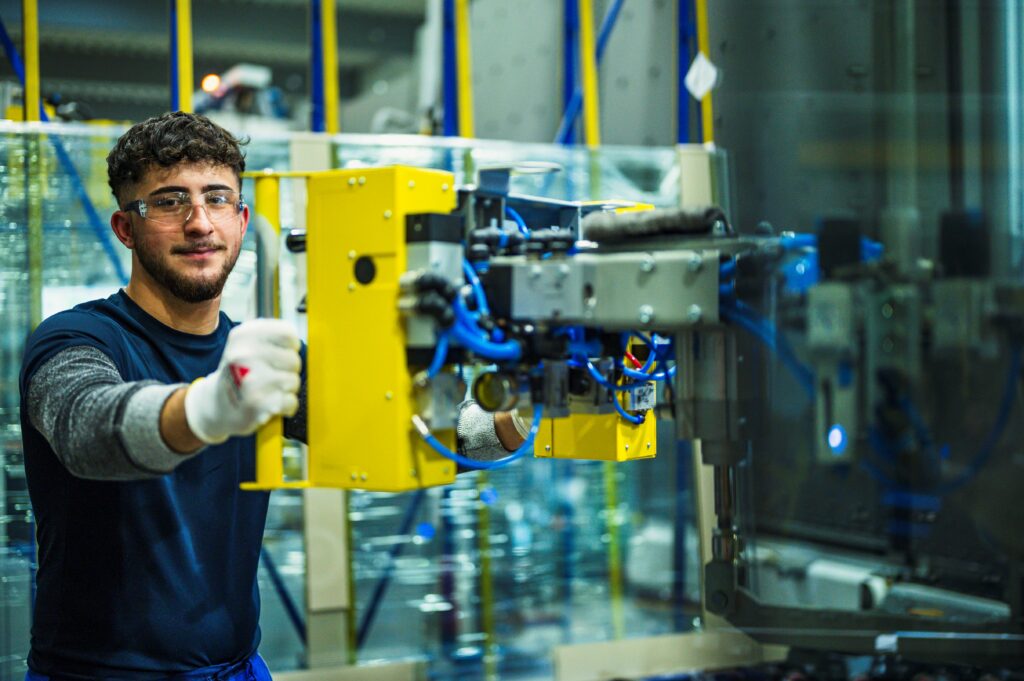 Ein junger Fabrikarbeiter bedient eine gelbe industrielle Maschine in einer modernen Produktionshalle. Er trägt Sicherheitshandschuhe und Schutzbrille und schaut freundlich in die Kamera.