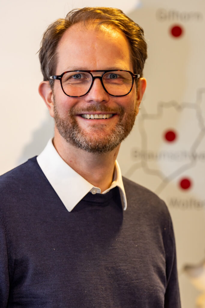 Ein Mann der direkt in die Kamera schaut und lächelt. Er trägt eine Brille und ein Hemd mit einem Pullover drüber.