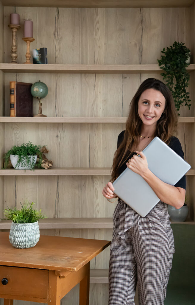 Es ist eine Frau zu sehen, die einen Laptop in der Hand hält. Sie steht vor einem Regal, welches mit einigen Deko-Artikel gefüllt ist. Die Frau lächelt in die Kamera. Sie hat braune lange Haare, ein schwarzes Oberteil an und trägt eine karierte Hose.