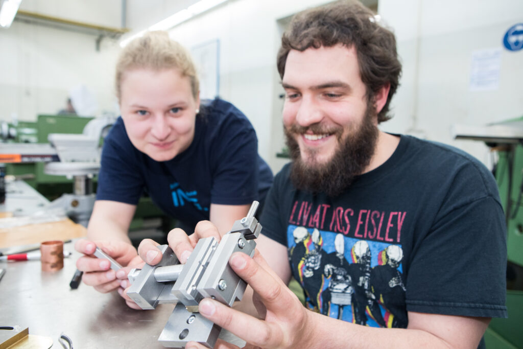 Es sind zwei junge Leute zu sehen, die einen metallischen Gegenstand in den Händen halten. Sie gucken es sich interessiert an.
