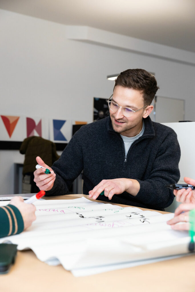 Junger Mann sitzt ein einem Tisch und hält einen Marker in der Hand. Er schaut auf das Poster vor ihm.
