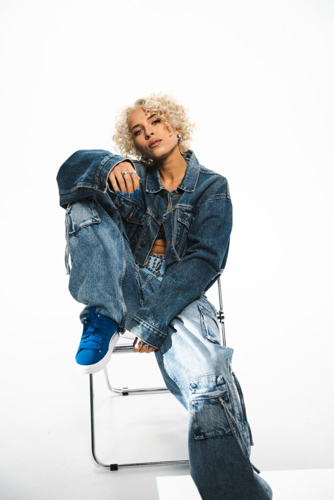 Künstlerin Zavet sitzt auf einem durchsichtigem Plastikstuhl. Ihr eines Bein ist angewinkelt und ihr Fuß steht auf dem Stuhl. Sie trägt eine Jeansjacke, Jeanshose und blaue Sneaker. 